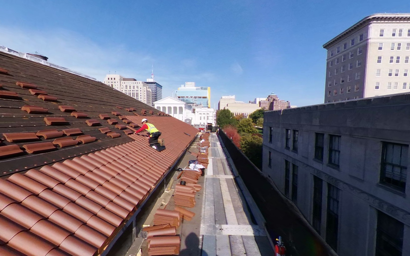 Photo of a worker installing a new tile roof on the Lewis F. Powell, Jr. courthouse. There are four rows of red tile shingles installed in the foreground near the roof edge, with more rows in the background. The buildings of downtown Richmond are visible in the background.